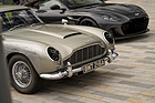 Aston Martin DB5 (2020) - Filmauto aus "Keine Zeit zu sterben" mit Gatling-Guns und Kampfspuren, dahinter ein moderner DBS