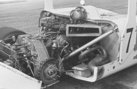 Chaparral 2H (1969) – Version mit hohem Flügel. Über der Hinterachse das wuchtige De-Dion-Rohr in Kastenform, welches die beiden Achsschenkel starr verbindet. Links unten ein Hydraulikzylinder, um den Bodenabstand bei verschiedenen Anpressdrücken konstant zu halten. Darüber ein kurzer Längslenker zur Längsführung des De-Dion-Rohrs, ein weiterer sichtbar in dessen Mitte, angelenkt am linken Arm des Rohrs. Darüber U-förmiger liegender Stabilisator, links ausgehängt. Direkt am Motor angelenkt das rechte Federbein. Massiver Schräglenker zwischen Kunststoff-Monocoque und Motor-/Hinterachs- Zusatzchassis (nicht sichtbar). Auspuffrohr seitlich austretend.