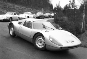 Einer der Porsche-904-Prototypen – erkennbar an dem seitlichen Lufteinlass ohne Hutze – in der Einfahrt zum neuen mehrstöckigen Porsche-Hauptquartier vor einer stattlichen Reihe an Porsche 356