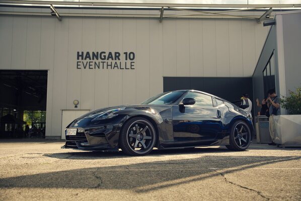 Nissan 370Z (2012) — The front bumper and forged wheels almost blend into the black paint; behind them is the facade of the Hangar 10 event hall